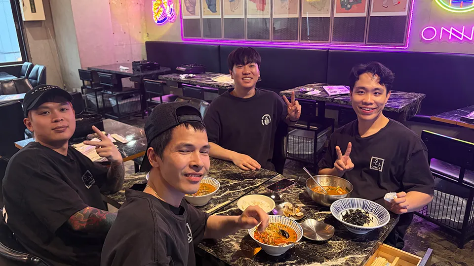 ネオンが光る店内で若い男性四人が食事を囲み、笑顔でピースサインをしている。