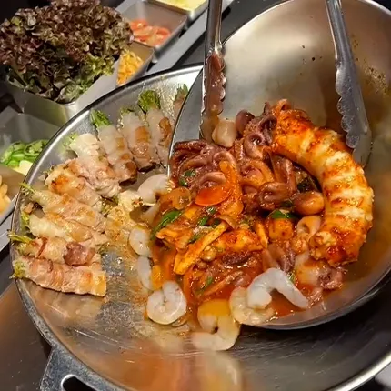 大きな金属鍋に辛い海鮮と焼き豚が盛られ、食欲をそそる彩り豊かな一品です